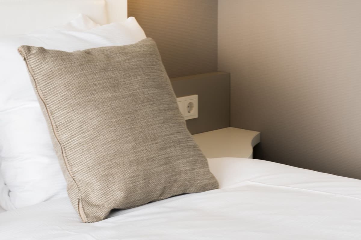 Uma cama bem arrumada com lençóis brancos e travesseiro bege, uma mesa de cabeceira e uma luminária fixada na parede.