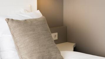 A neatly made bed with white linens and a beige pillow, a bedside table, and a wall-mounted lamp.