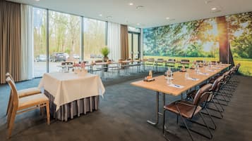 Un salón de conferencias con una larga mesa, sillas y un amplio ventanal con vistas a árboles y coches aparcados.