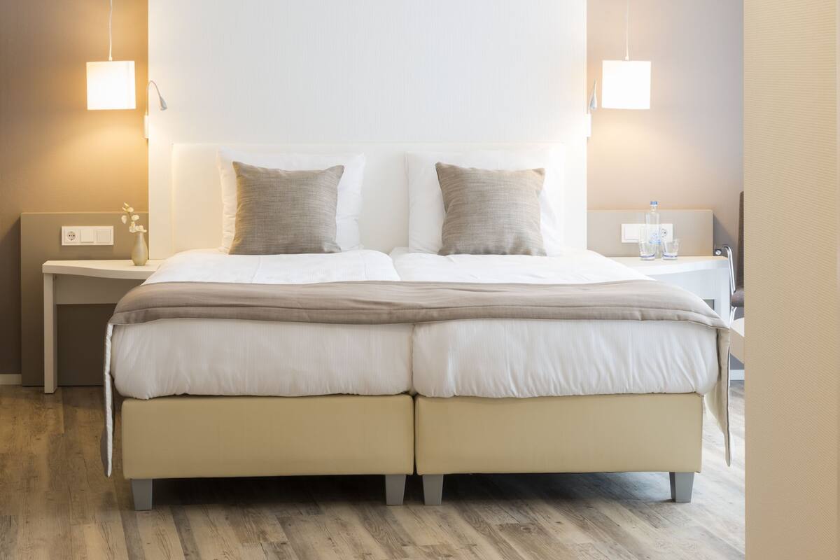 Uma cama de casal com cabeceira e peseira bege, duas almofadas e uma mesa de cabeceira com abajur.
