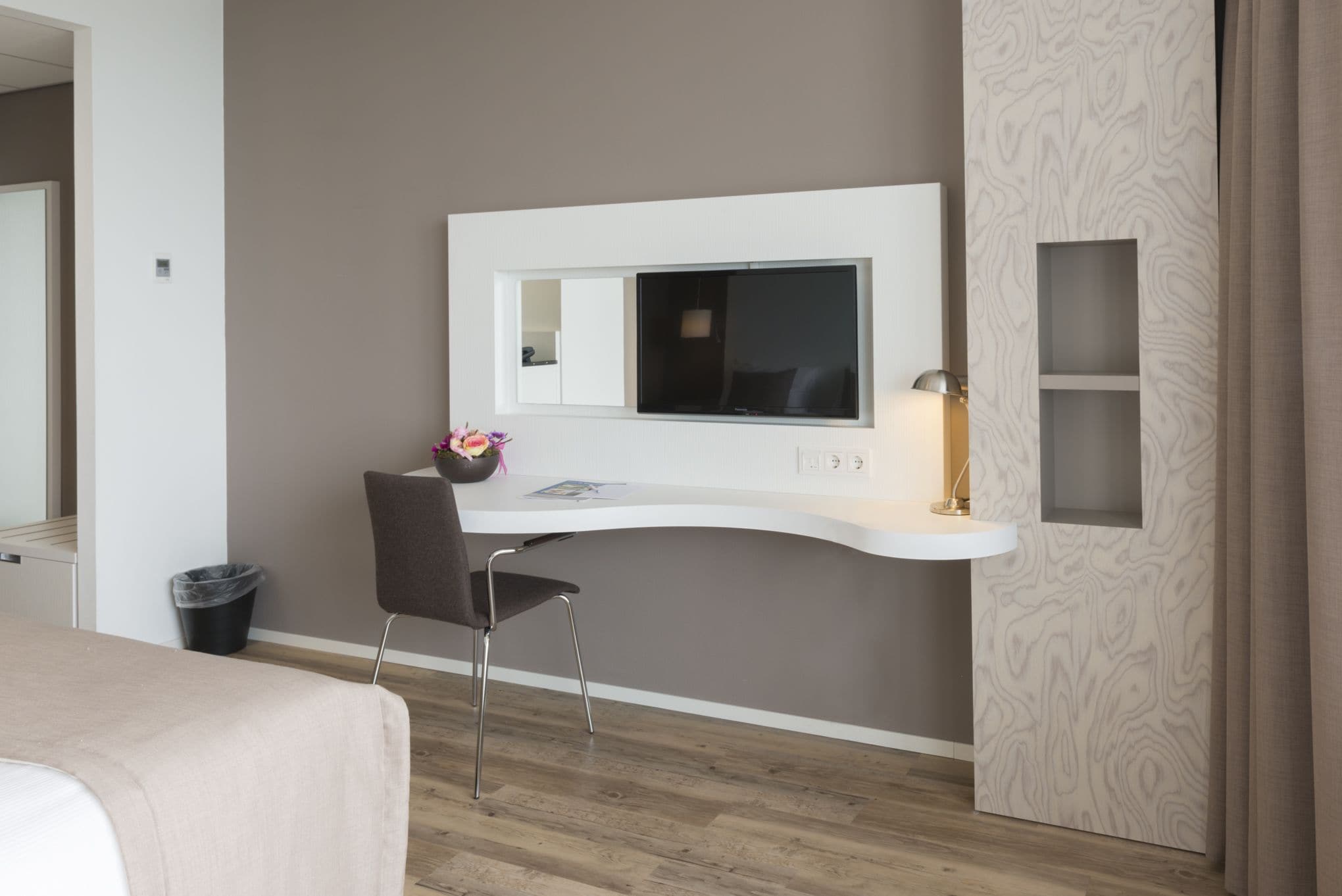 A modern hotel room with a curved desk, a television mounted on the wall, a small seating area with a chair, and a trash bin.