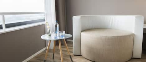 Una habitación moderna con un sillón redondo color beige, una mesita redonda con una botella de agua y vasos, y una ventana con cortinas.