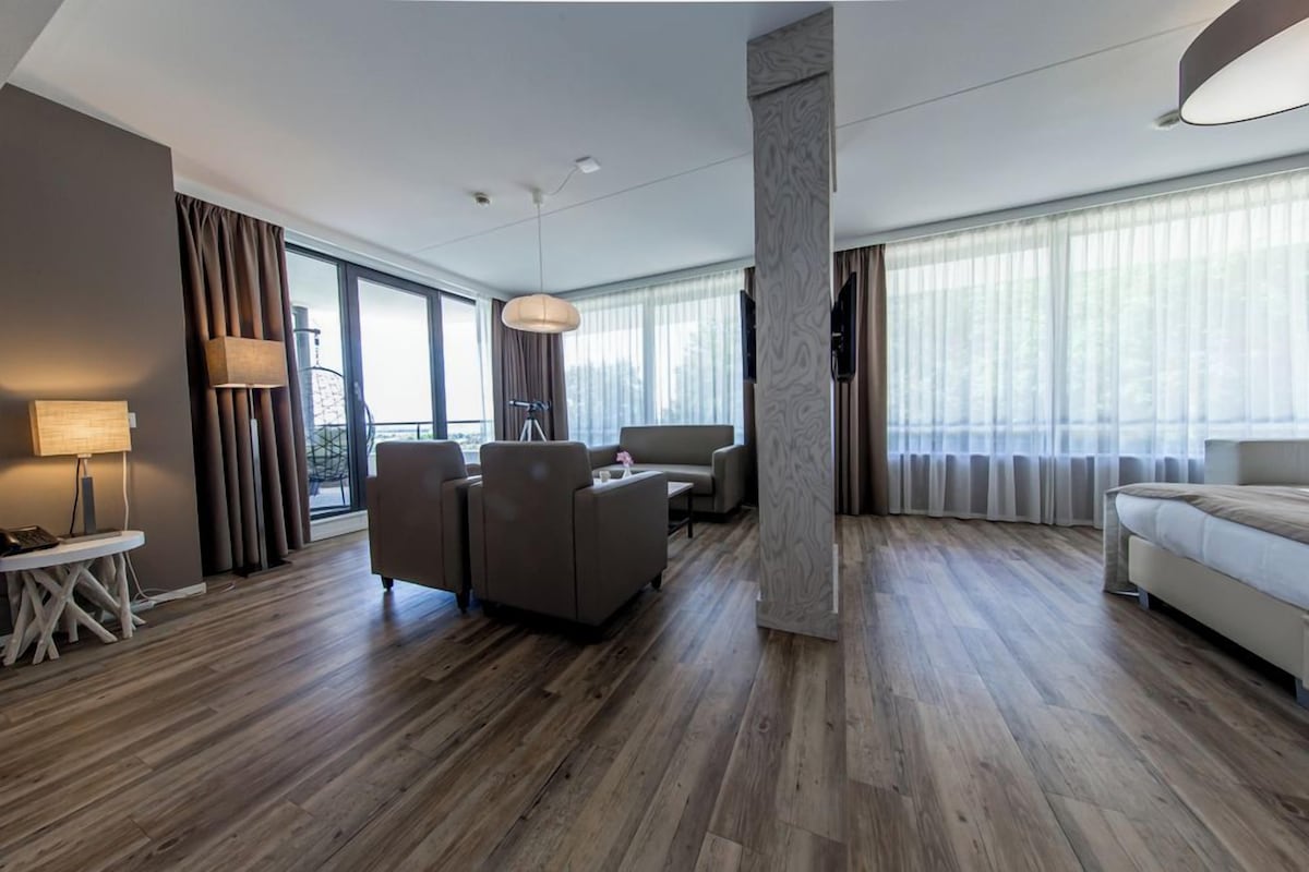 A modern hotel room with a wooden floor, a sofa, a coffee table, a lamp, and large windows with curtains.