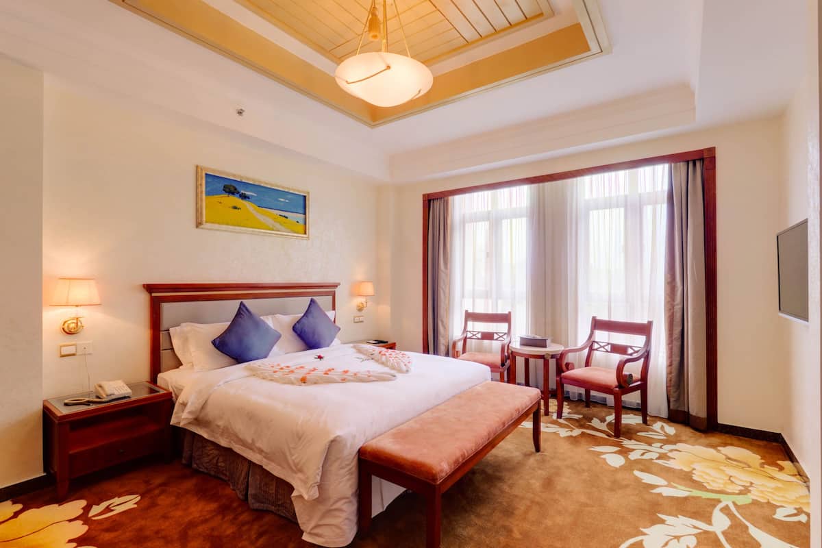 A hotel room with a large bed, two bedside tables, a desk with chairs, a painting on the wall, and a large window with curtains.