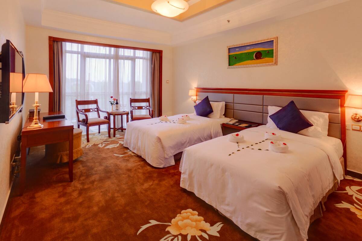 A hotel room with two beds, a desk with a lamp, a chair, a painting on the wall, and a large window with curtains.
