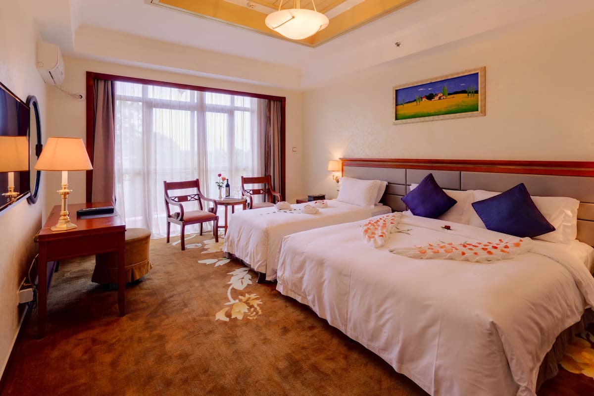 A hotel room with two beds, a desk with a lamp, a chair, a small table with a vase and wine glass, and a painting on the wall.