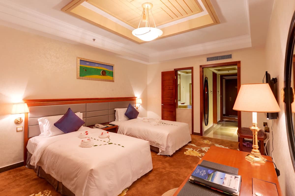 A hotel room with two beds, a desk, a lamp, a painting on the wall, and a door leading to another room.