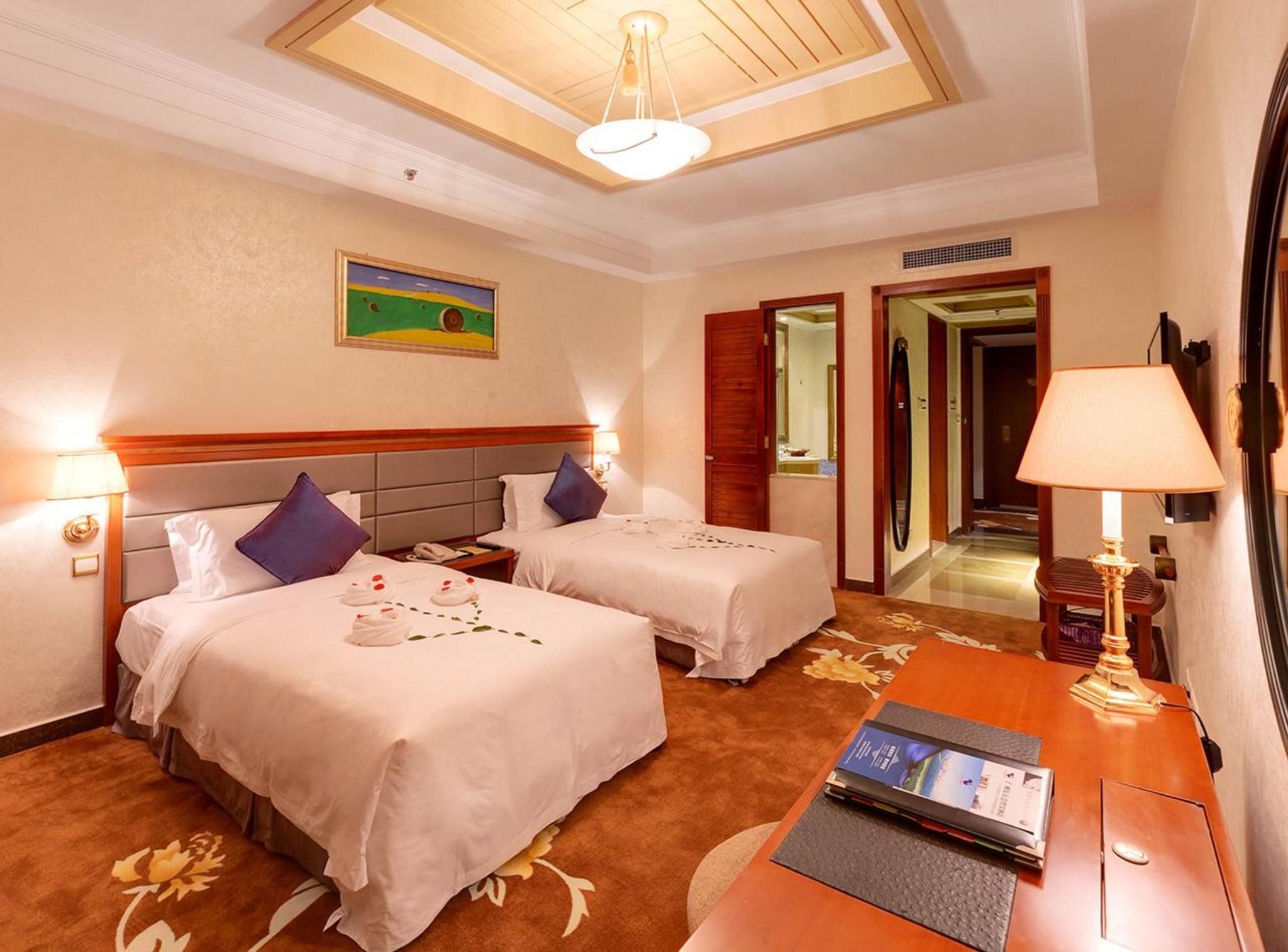A hotel room with two beds, a desk, a lamp, a painting, and a door leading to a hallway.