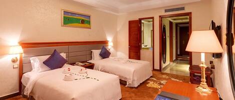A hotel room with two beds, a desk, a lamp, a painting, and a door leading to a hallway.