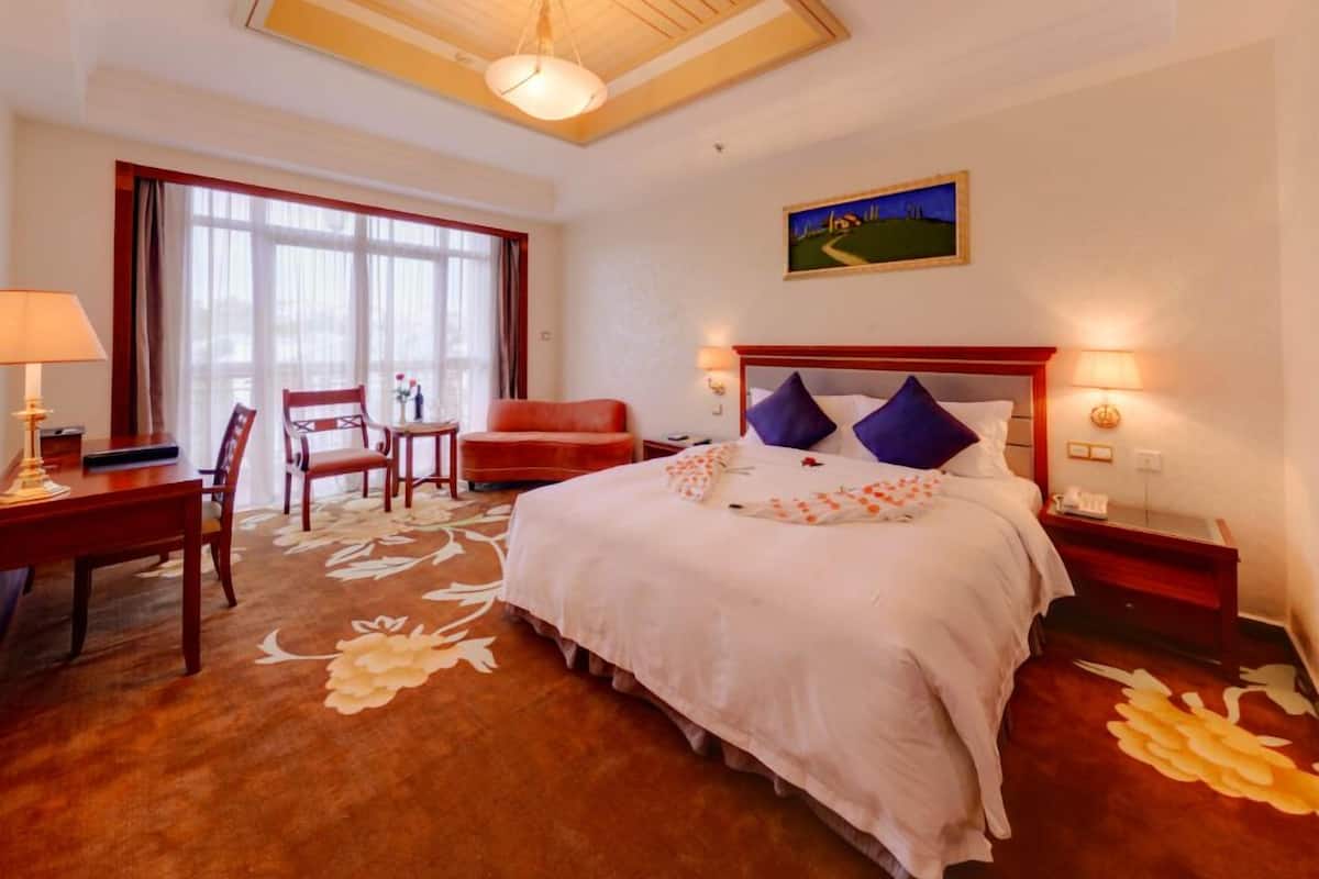 A hotel room with a large bed, a desk with a lamp, a chair, and a window with curtains.