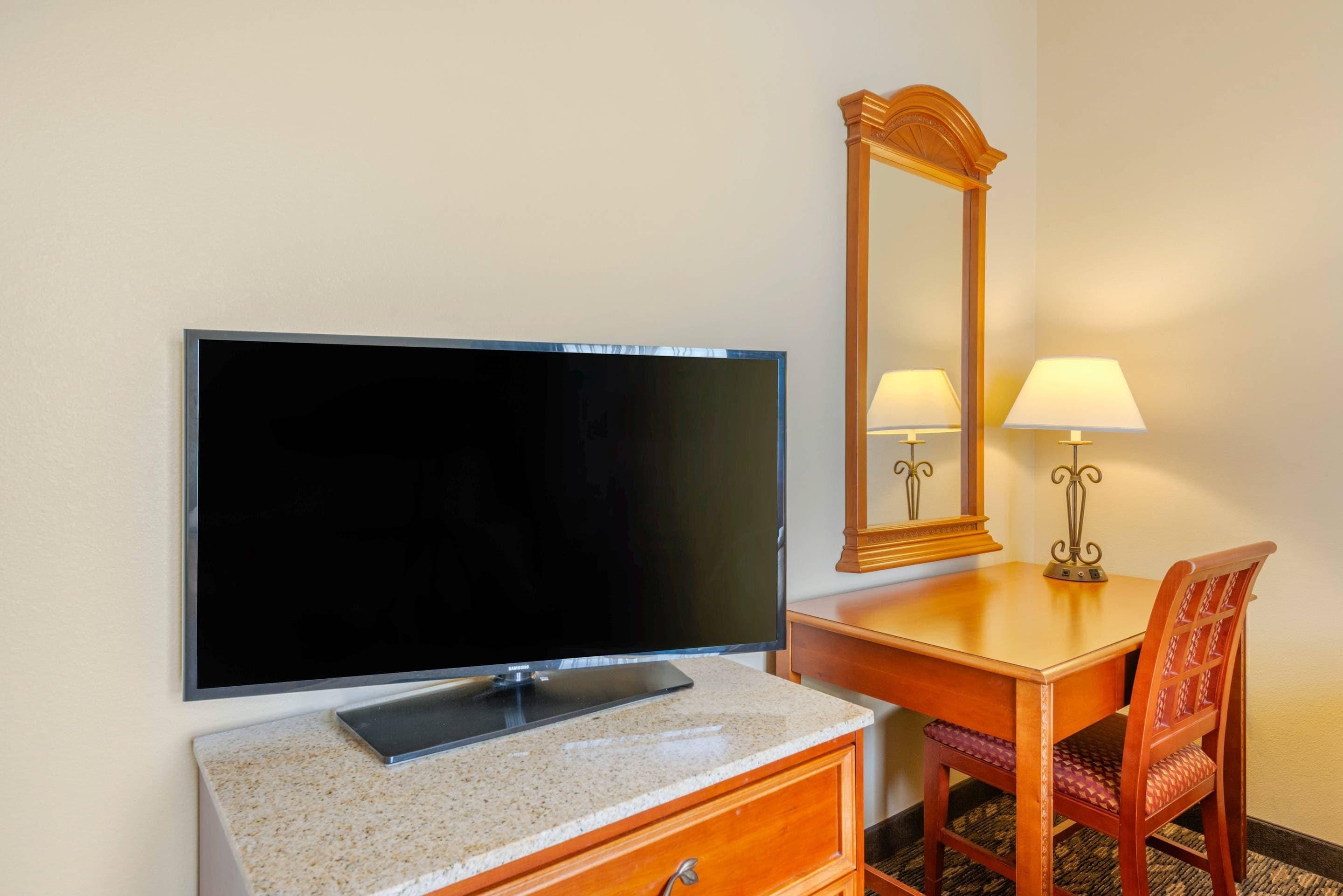 Ett hotellrum med en platt-TV som är monterad på väggen, ett skrivbord i trä med en lampa och en stol.