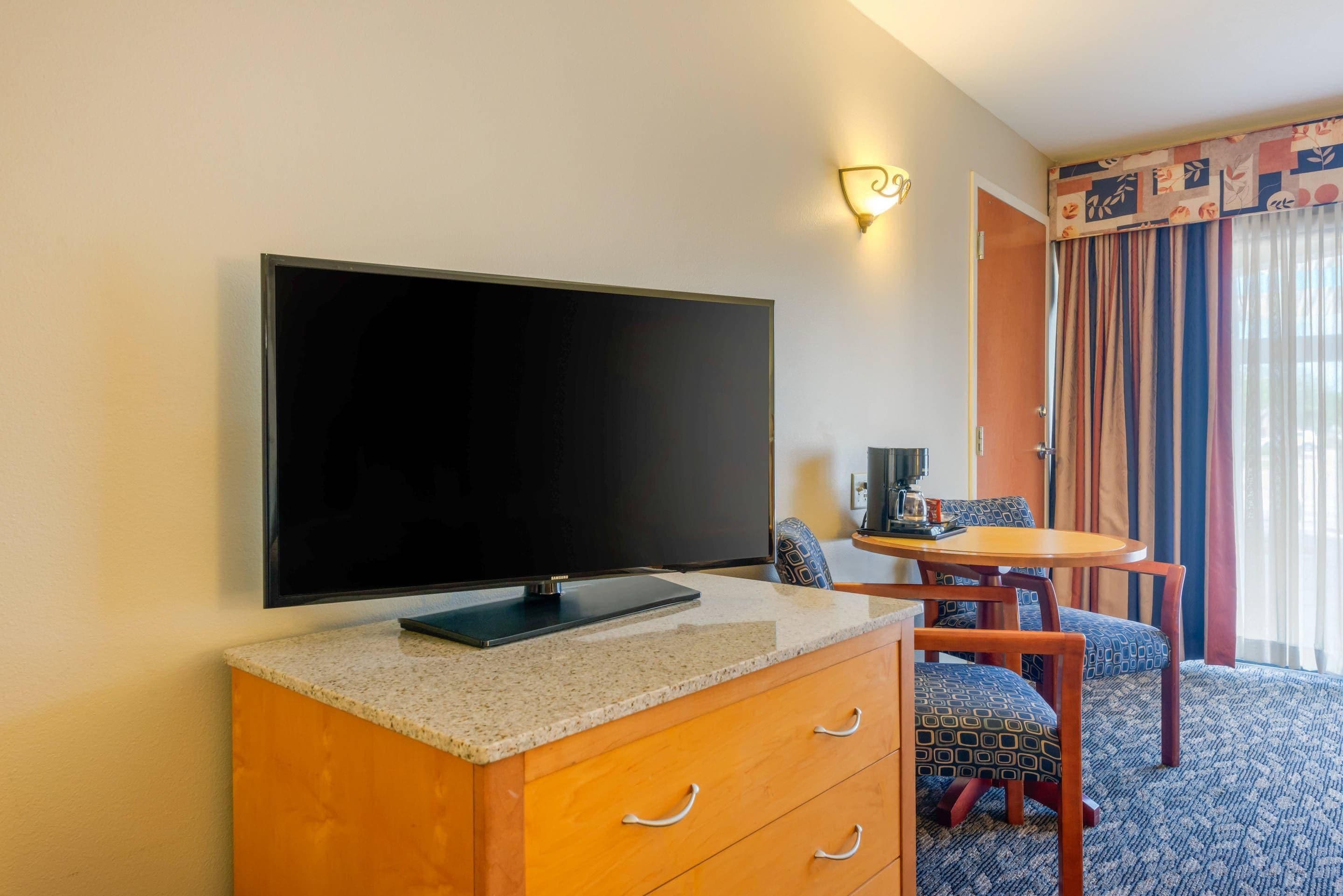 Une chambre d’hôtel avec un téléviseur à écran plat posé sur un meuble en bois, une petite table avec une machine à café, des chaises à motifs et une fenêtre avec des rideaux.