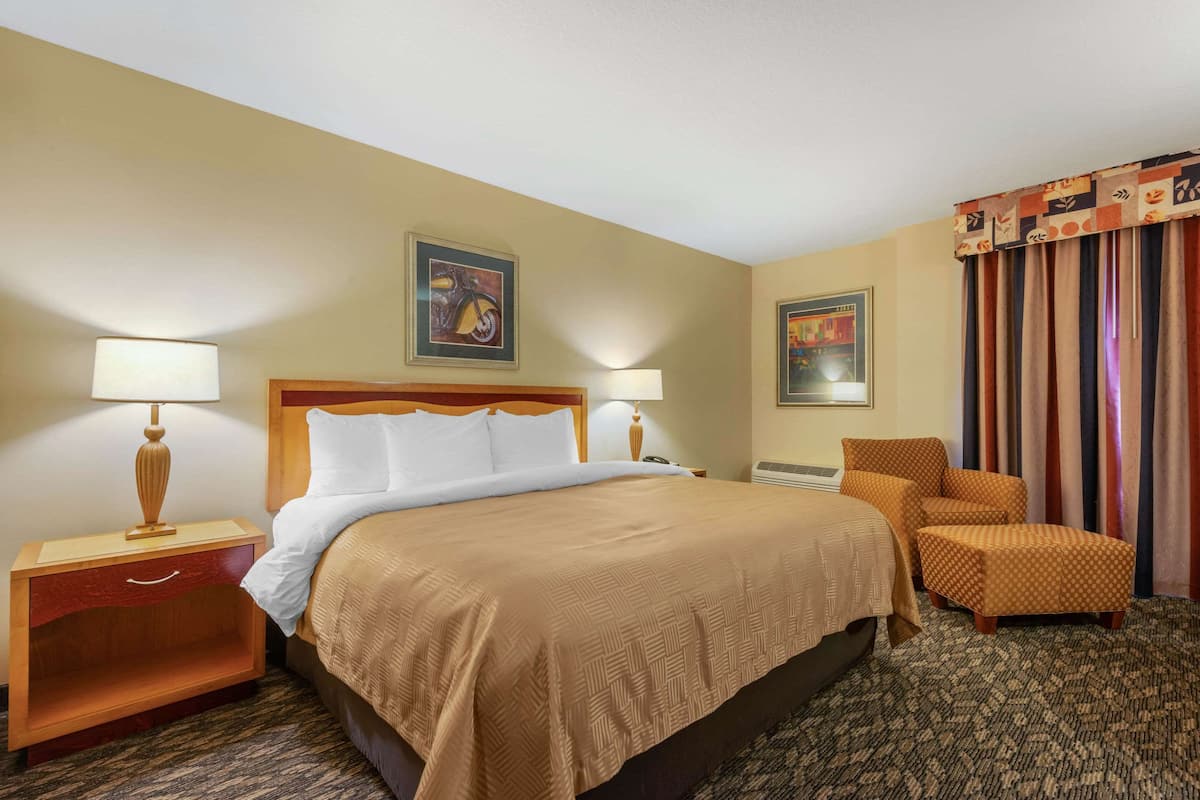 Una habitación de hotel con una cama grande, dos lámparas de noche, una mesita de noche con cajón, dos sillones y suelo alfombrado.