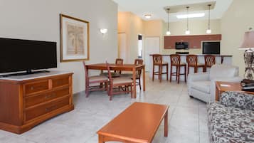Un soggiorno con televisore, arredi in legno, zona pranzo con sedie e cucina con forno a microonde.