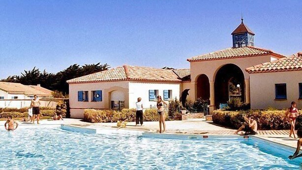 Une zone avec une piscine où se trouvent des personnes, un bâtiment avec un toit en tuiles et un clocher.