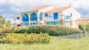 Un grand immeuble à plusieurs étages, doté d’une façade blanche et d’un toit recouvert de tuiles rouges. Il présente de nombreux balcons avec des garde-corps bleus et des fenêtres en arc de cercle.