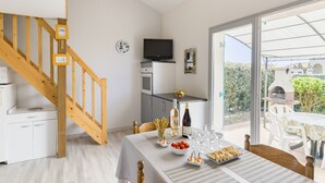 Une cuisine moderne avec un escalier en bois, un coin repas avec une table dressée pour deux personnes, et une vue sur le jardin à travers une porte vitrée.