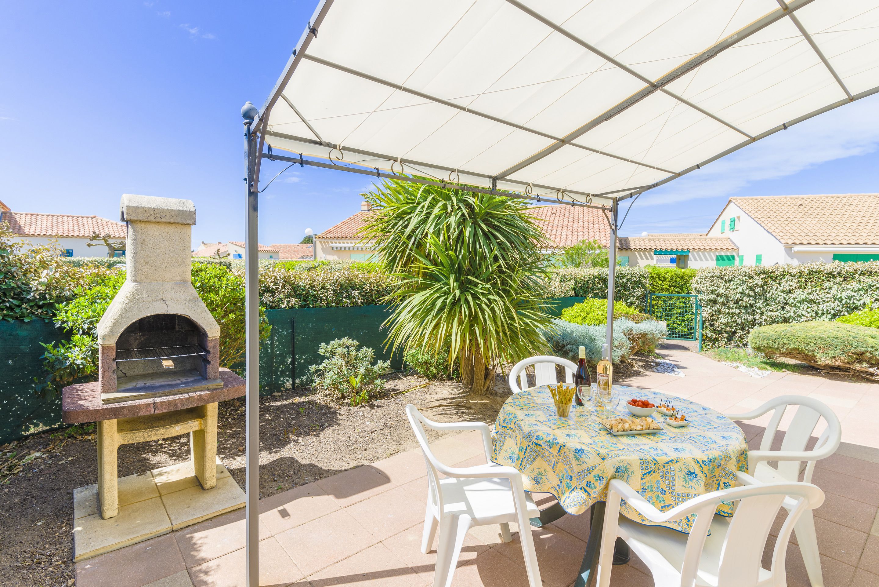 An outdoor patio with a stone-built pizza oven, white plastic chairs, and a table with a patterned tablecloth.