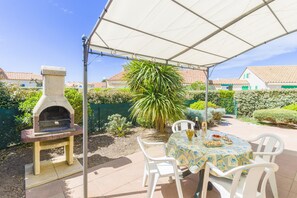 Een terras buiten, met een pizzaoven van natuursteen, witte plastic stoelen en een tafel met een tafelkleed met patroon.