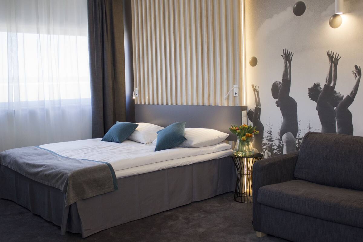 Une chambre d’hôtel moderne avec un grand lit, un canapé gris, une lampe fixée au mur et un élément décoratif mural.