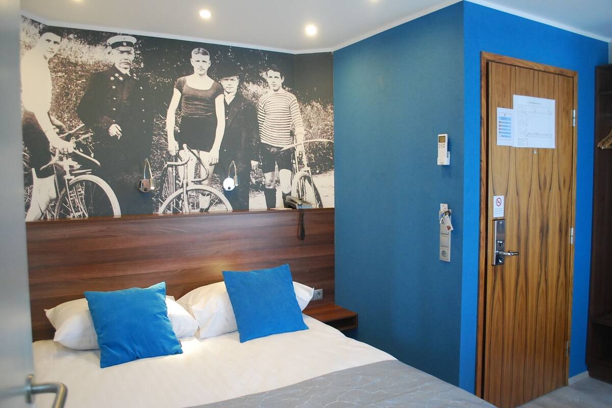 Une chambre d’hôtel avec un lit, des oreillers bleus, une tête de lit en bois, un mur orné d’une fresque photographique en noir et blanc, une porte avec une pancarte et un téléphone fixé au mur.
