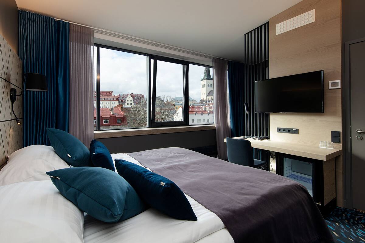 Une chambre d’hôtel avec un grand lit, un bureau doté d’un téléviseur à écran plat et une vue sur le paysage urbain par la fenêtre.