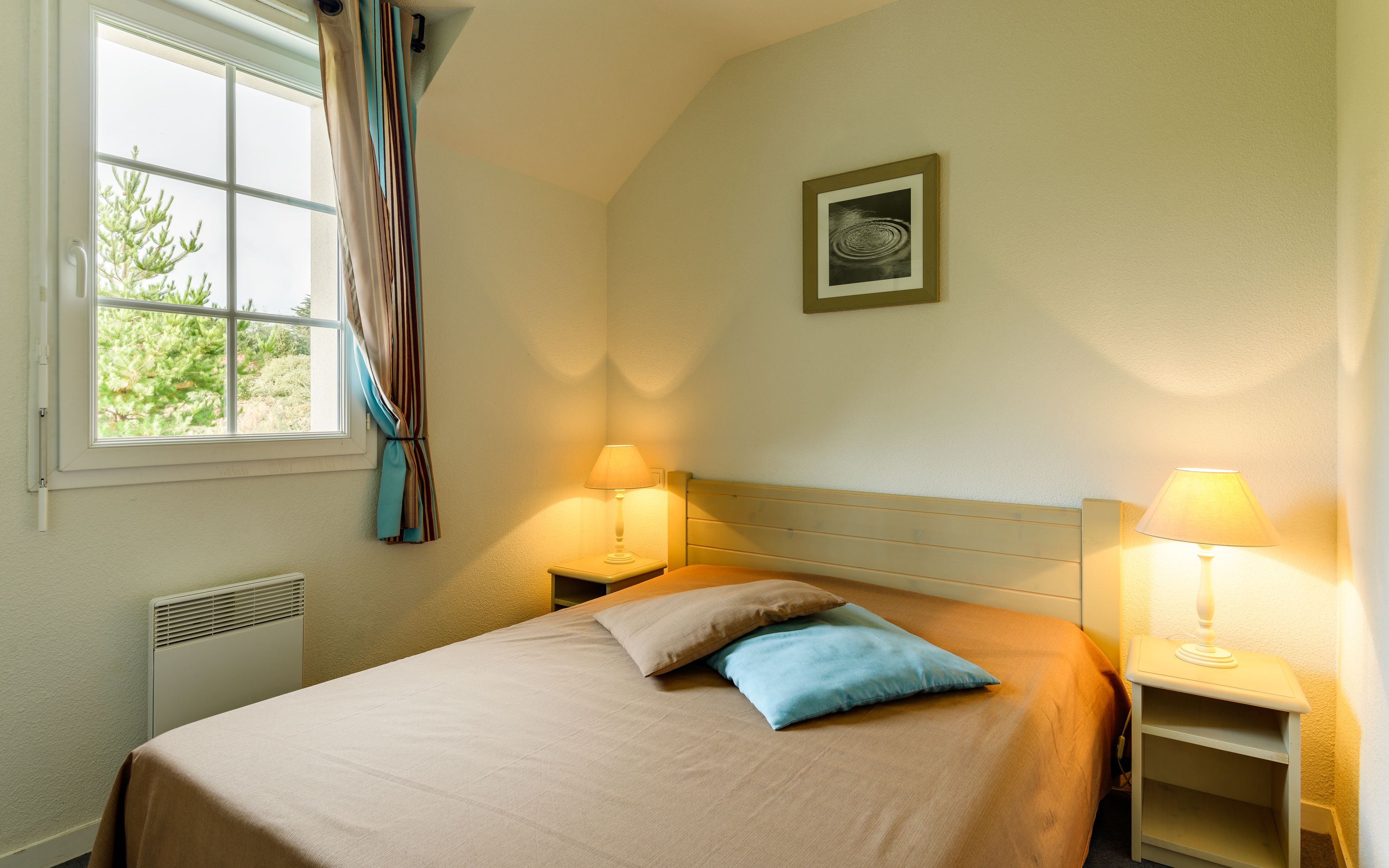 Un dormitorio con cama, dos lámparas de noche, una ventana con cortinas, un radiador y un cuadro en la pared.
