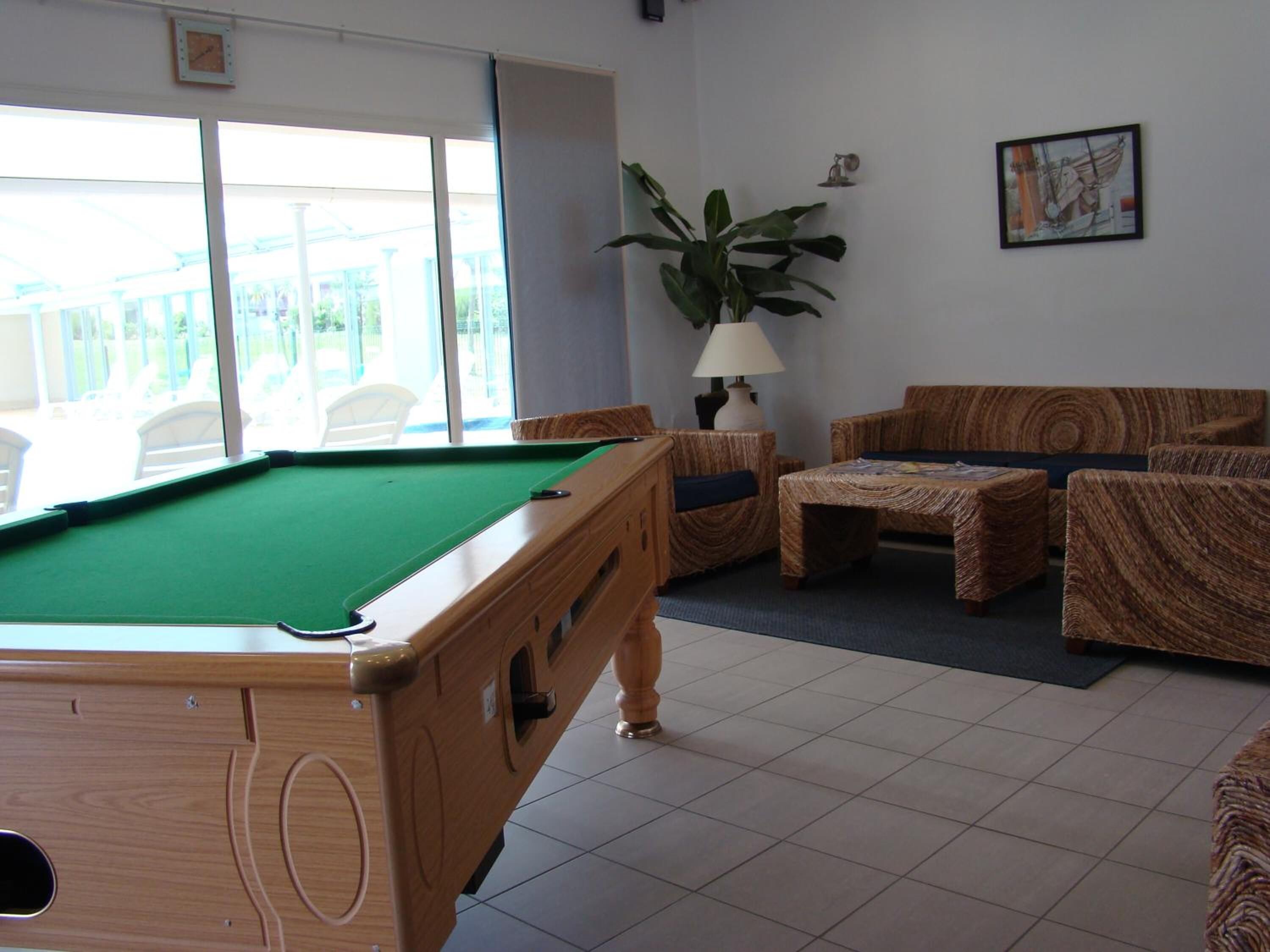 A pool table in a room with a view of a balcony. There are two wooden chairs and a small table with a lamp.