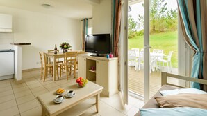A room with a dining table, chairs, a TV, a small kitchen area, and a sliding glass door leading to an outdoor patio with white furniture.