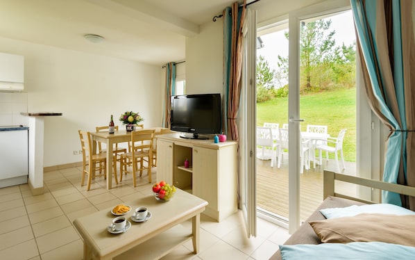 A room with a dining table, chairs, a TV, a small kitchen area, and a sliding glass door leading to an outdoor patio with white furniture. - Résidence Lagrange Vacances le Hameau de Peemor Pen (Crozon)