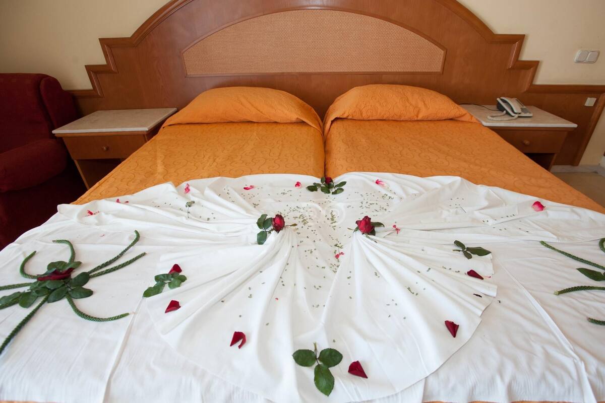 Un lit bien fait, recouvert d’une couverture orange et d’un couvre-lit blanc orné de pétales de roses et de feuilles vertes.
