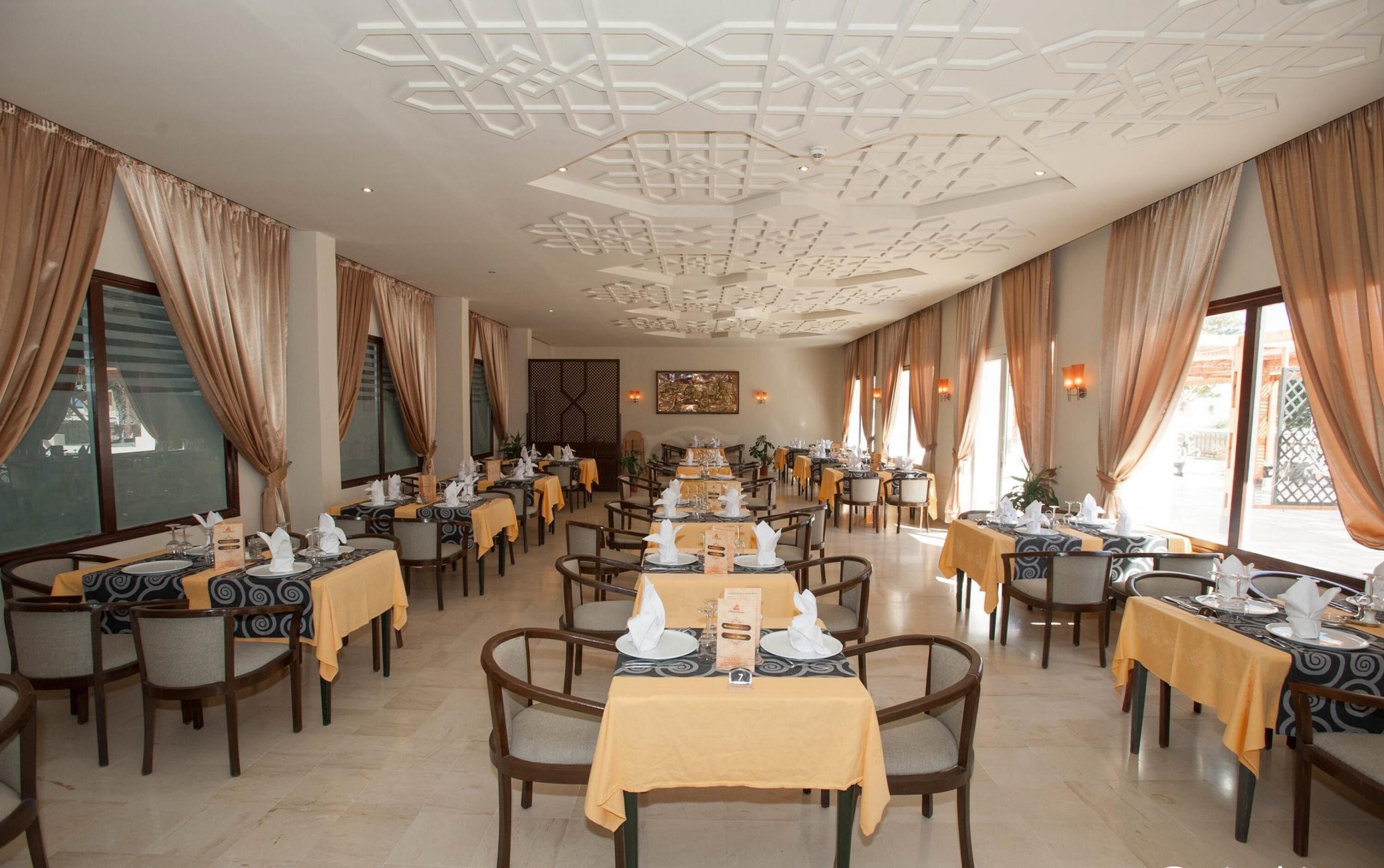 A restaurant with multiple tables, each set with plates, glasses, and napkins. The room has patterned ceiling and large windows with curtains.