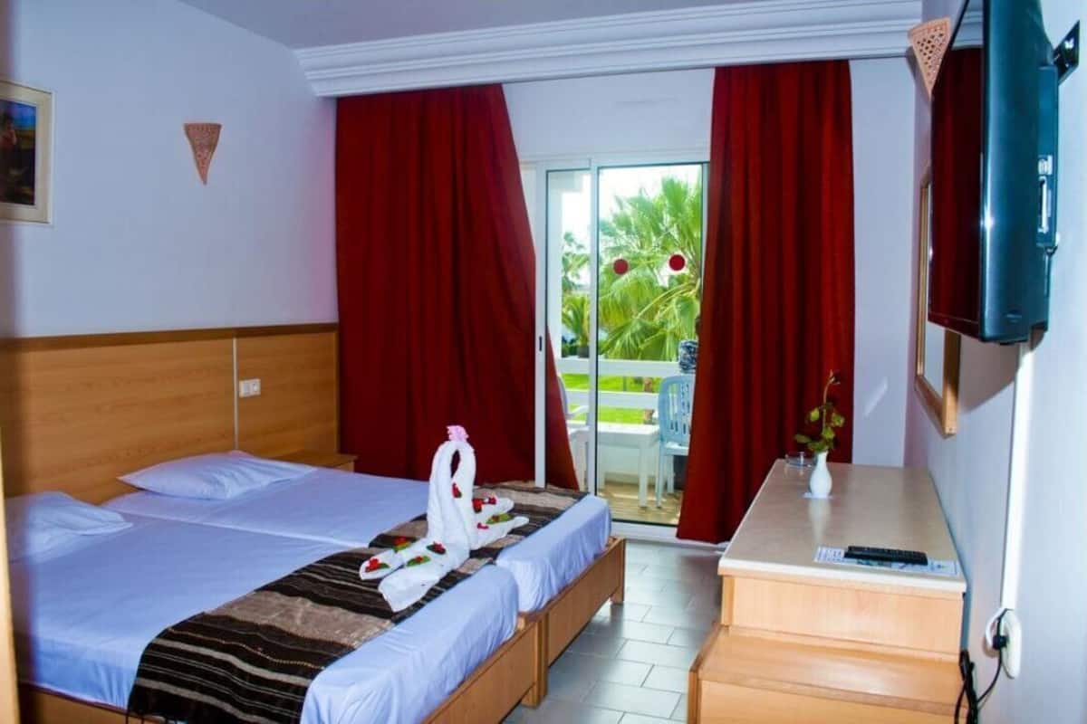 Une chambre d’hôtel avec deux lits, une tête de lit en bois, un petit bureau, une télévision et un balcon avec des rideaux rouges.