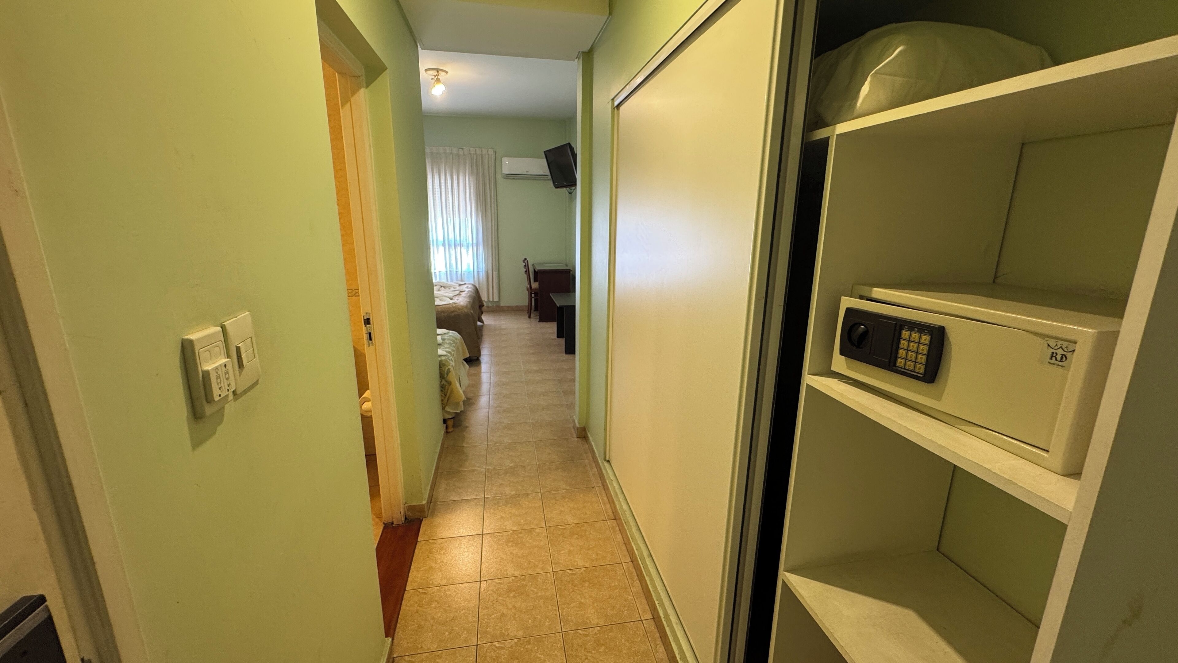 A narrow hallway with a door on the left, a bed with a white cover in the middle, and a white cabinet with a lock on the right.