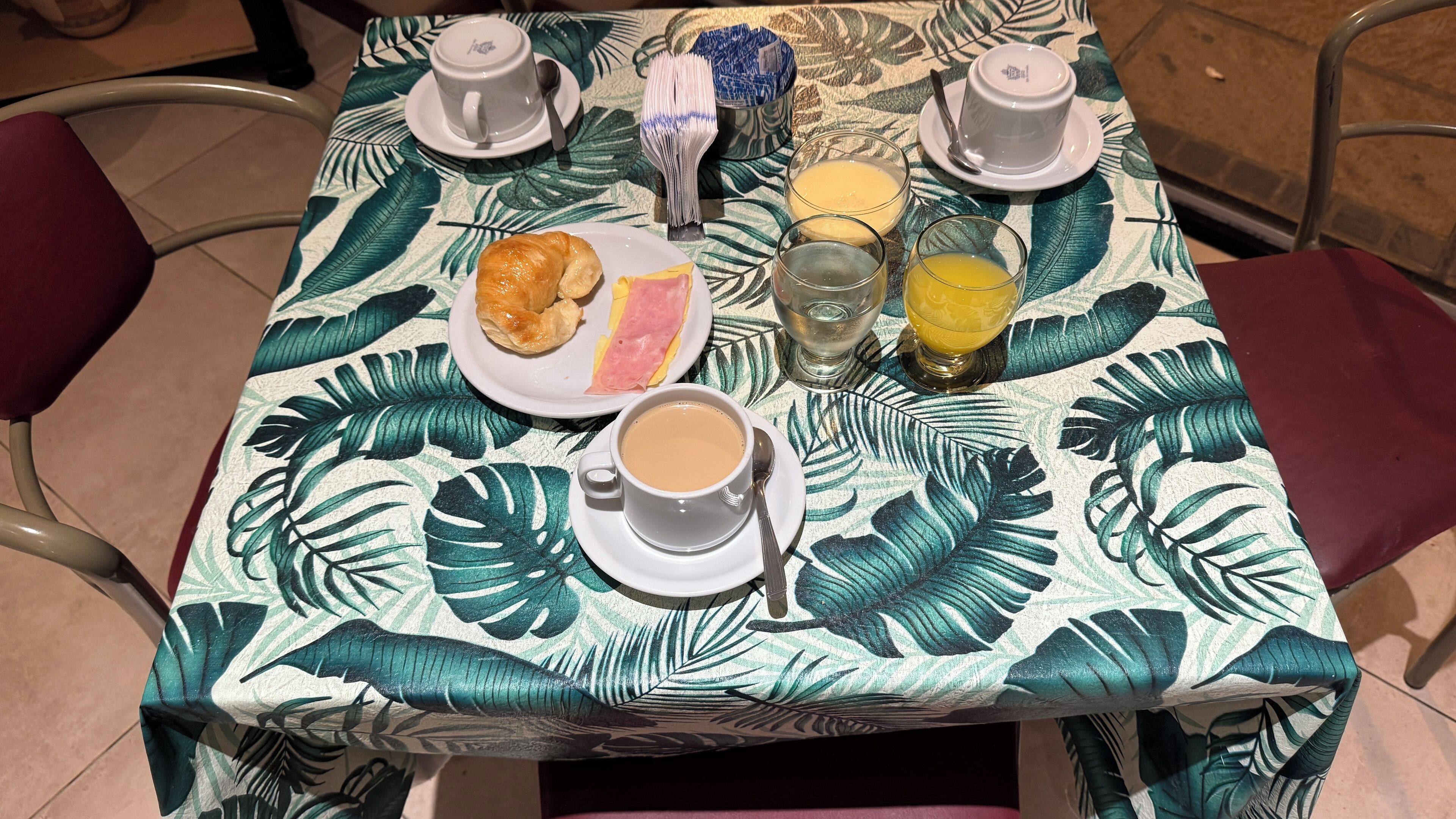 Una mesa con mantel de hojas tropicales, un croissant, una rebanada de pastel, un vaso de jugo de naranja, un vaso de agua y una taza de café.