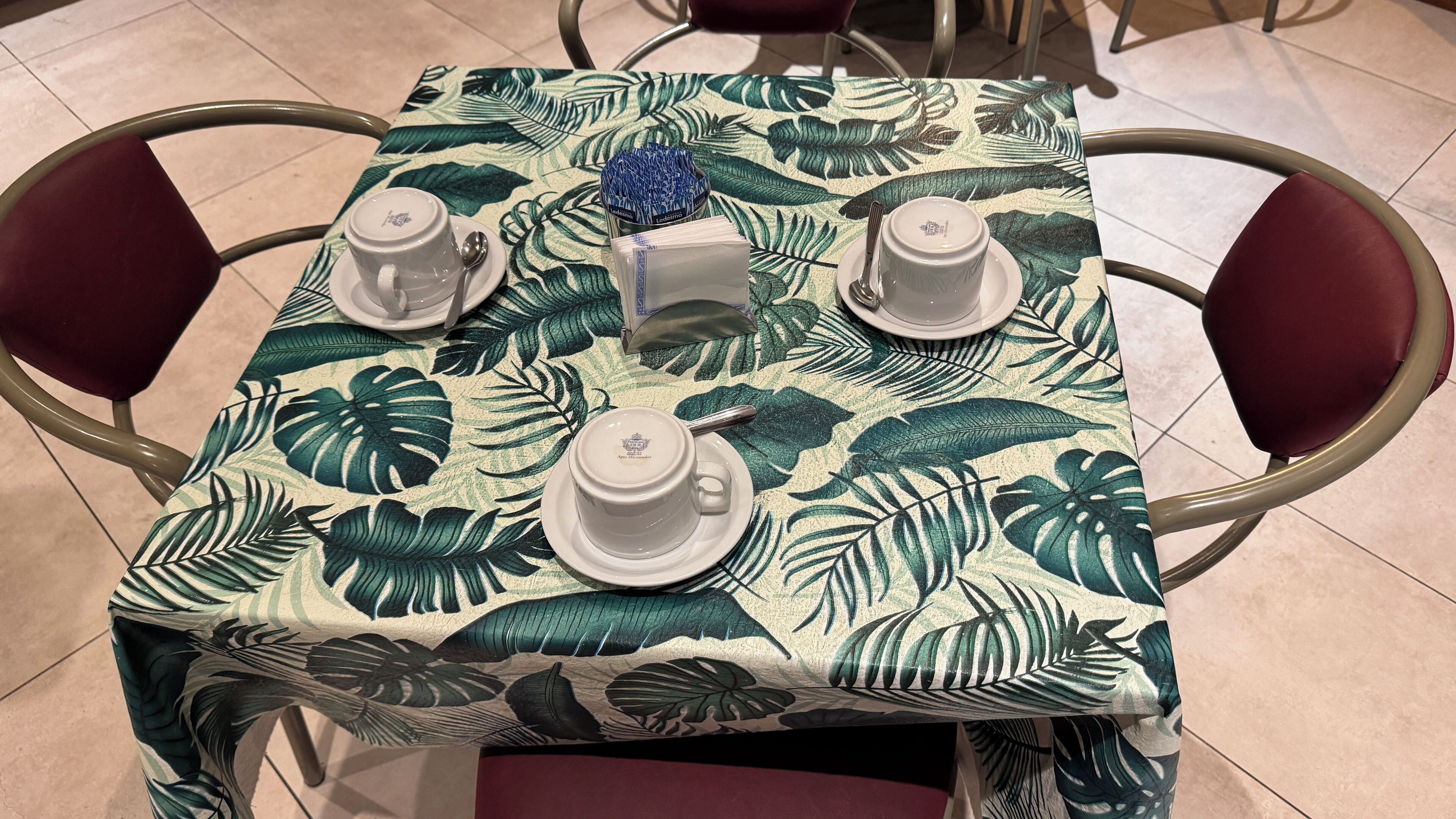 Ein quadratischer Tisch mit tropischem Blattmuster, vier weiße Tassen mit Untertellern, eine blaue Serviettenbox und ein kleines blaues Dekorationsstück.