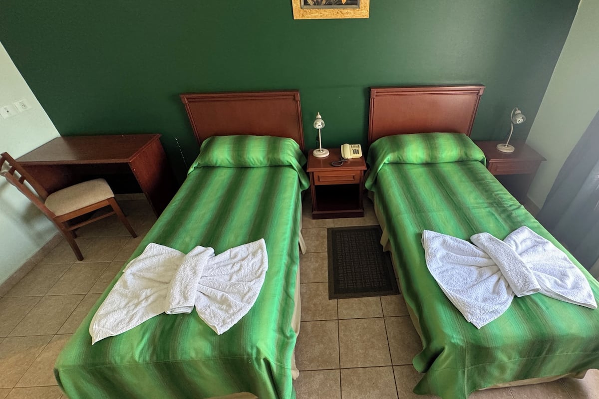 A room with two single beds, each with a green bedspread and white towels. There is a wooden nightstand with a telephone and a lamp between the beds. A wooden chair is placed next to a desk with a chair. The room has a green wall and a window with curtains.
