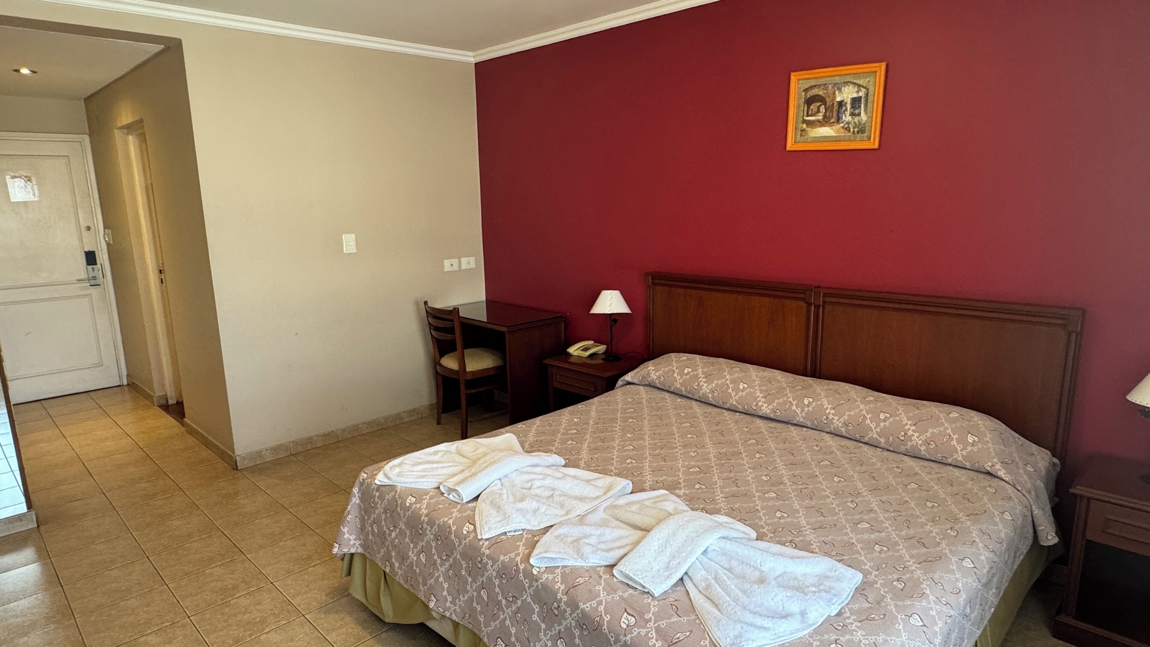 A hotel room with a bed, a chair, a desk, and a painting on the wall.