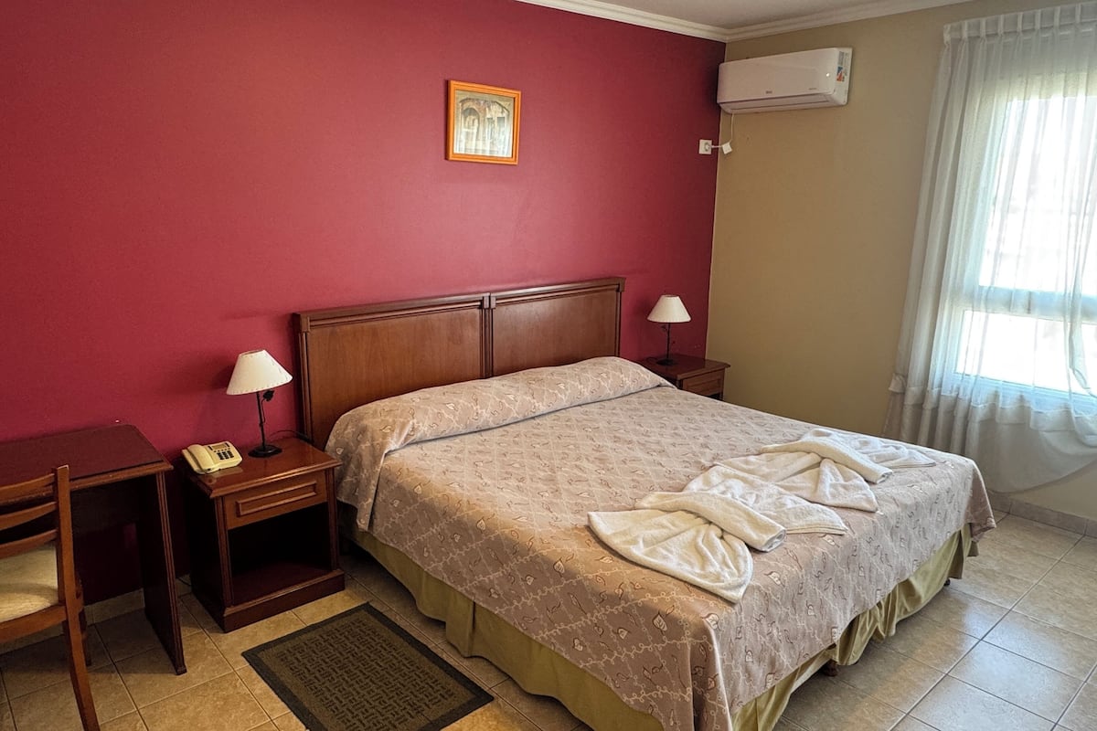 A hotel room with a bed, a desk, a chair, a telephone, and an air conditioning unit.