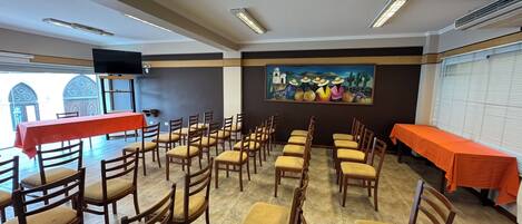 Una sala de conferencias con filas de sillas y mesas de madera, un cuadro en la pared y un televisor montado en el techo.
