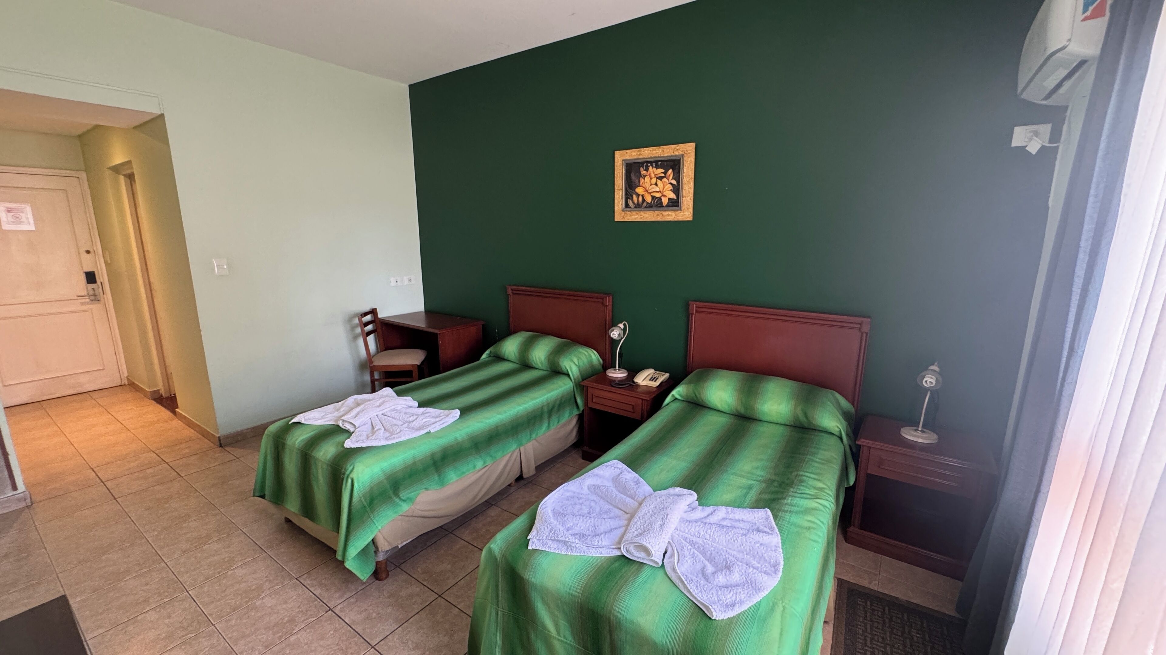 A hotel room with two single beds, a desk, a chair, a nightstand with a lamp, and a wall-mounted picture.