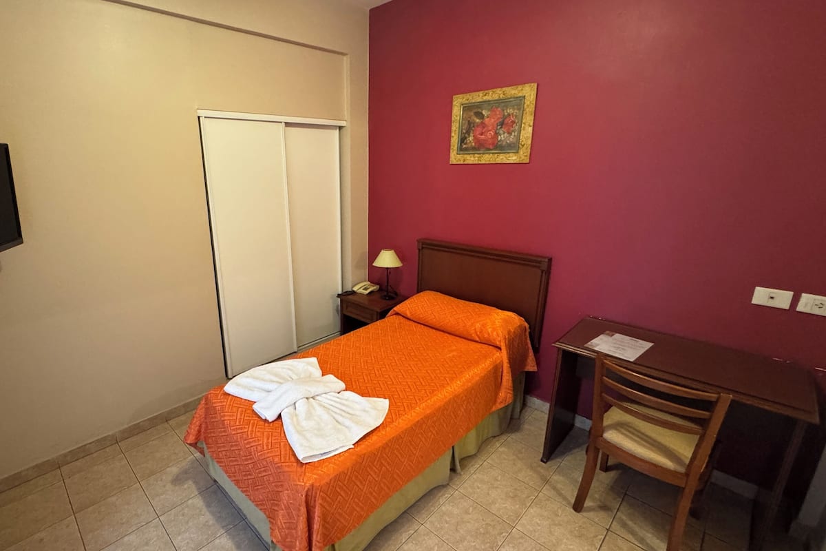A small, tidy room with a bed, a desk, a chair, a TV, and a door.