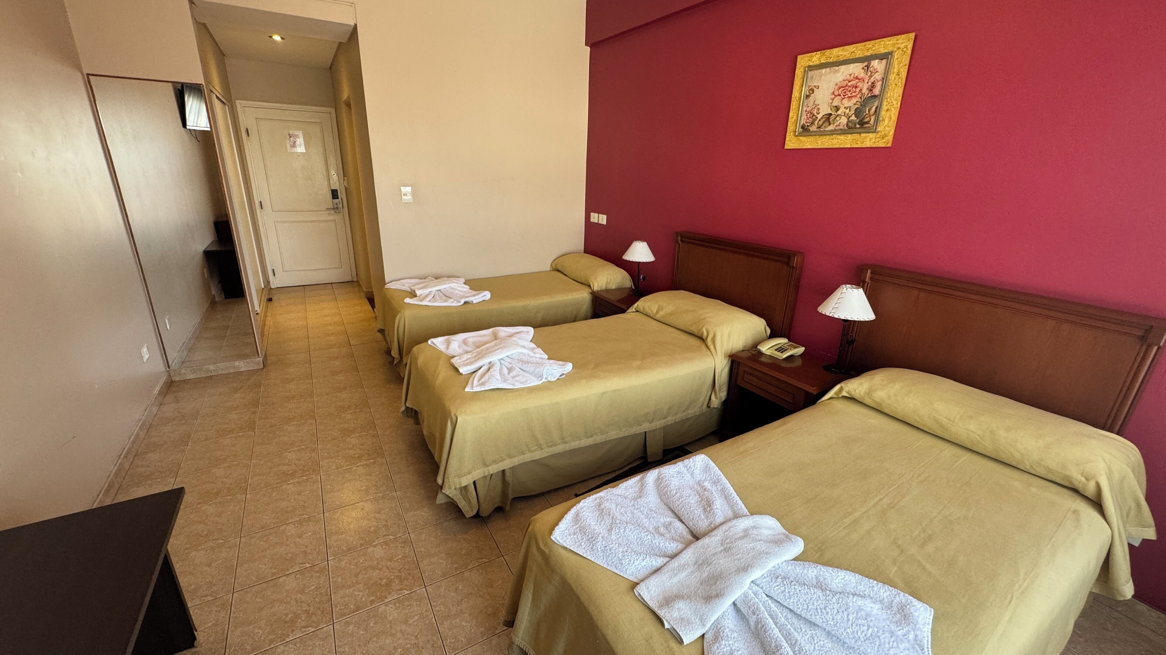 Ein Hotelzimmer mit vier Einzelbetten, auf jedem Bett ein weißes Handtuch. Rechts befindet sich ein Holz-Kopfteil, ein Nachttisch mit Lampe und Telefon, sowie eine rote Wand mit einem gerahmten Bild.