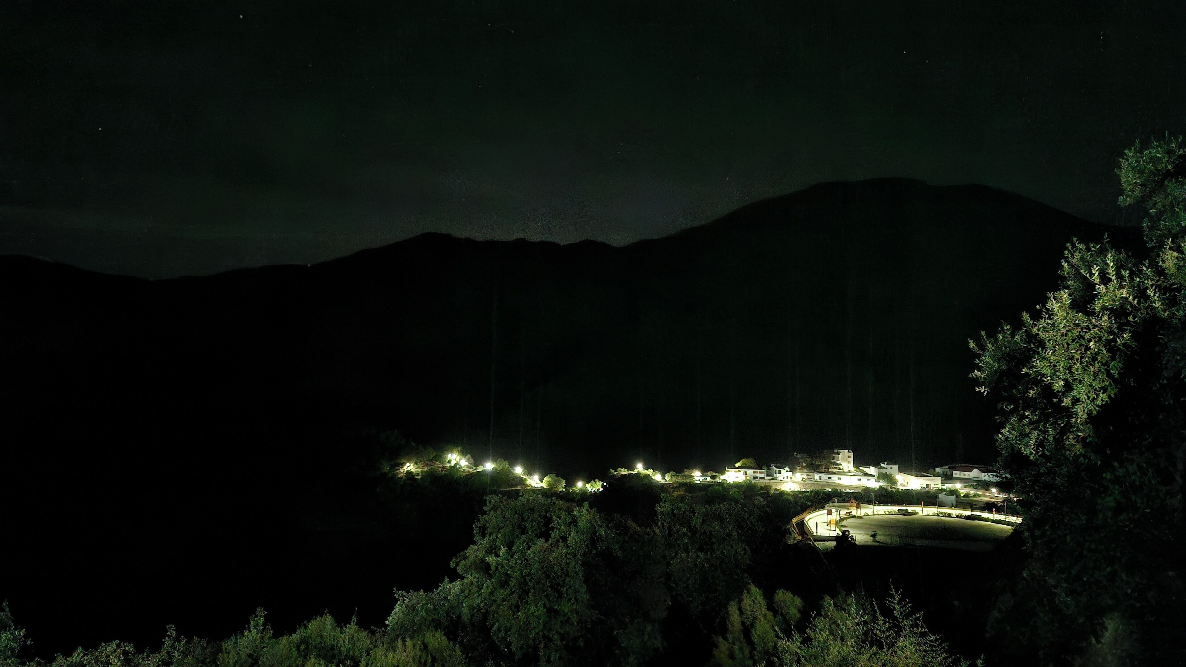 Une vue nocturne d’un village côtier, avec des bâtiments illuminés et une route sinueuse.