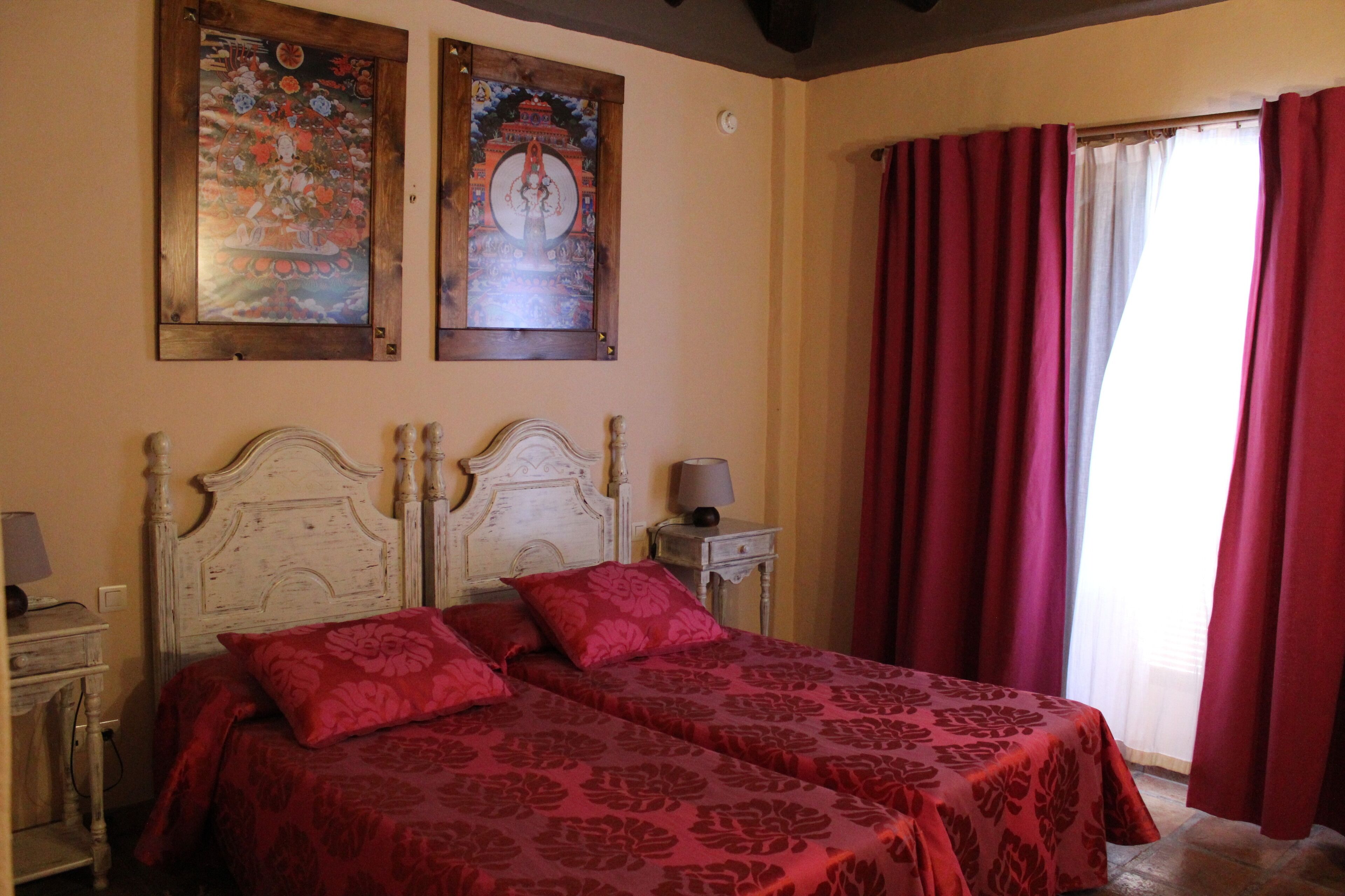 Uma cama de casal com roupa de cama estampada em flor vermelha, uma mesa de cabeceira com abajur e duas obras de arte emolduradas na parede.