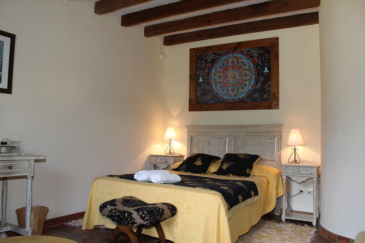 A bedroom with a bed, two bedside tables, a lamp, a chair, and a decorative wall hanging.