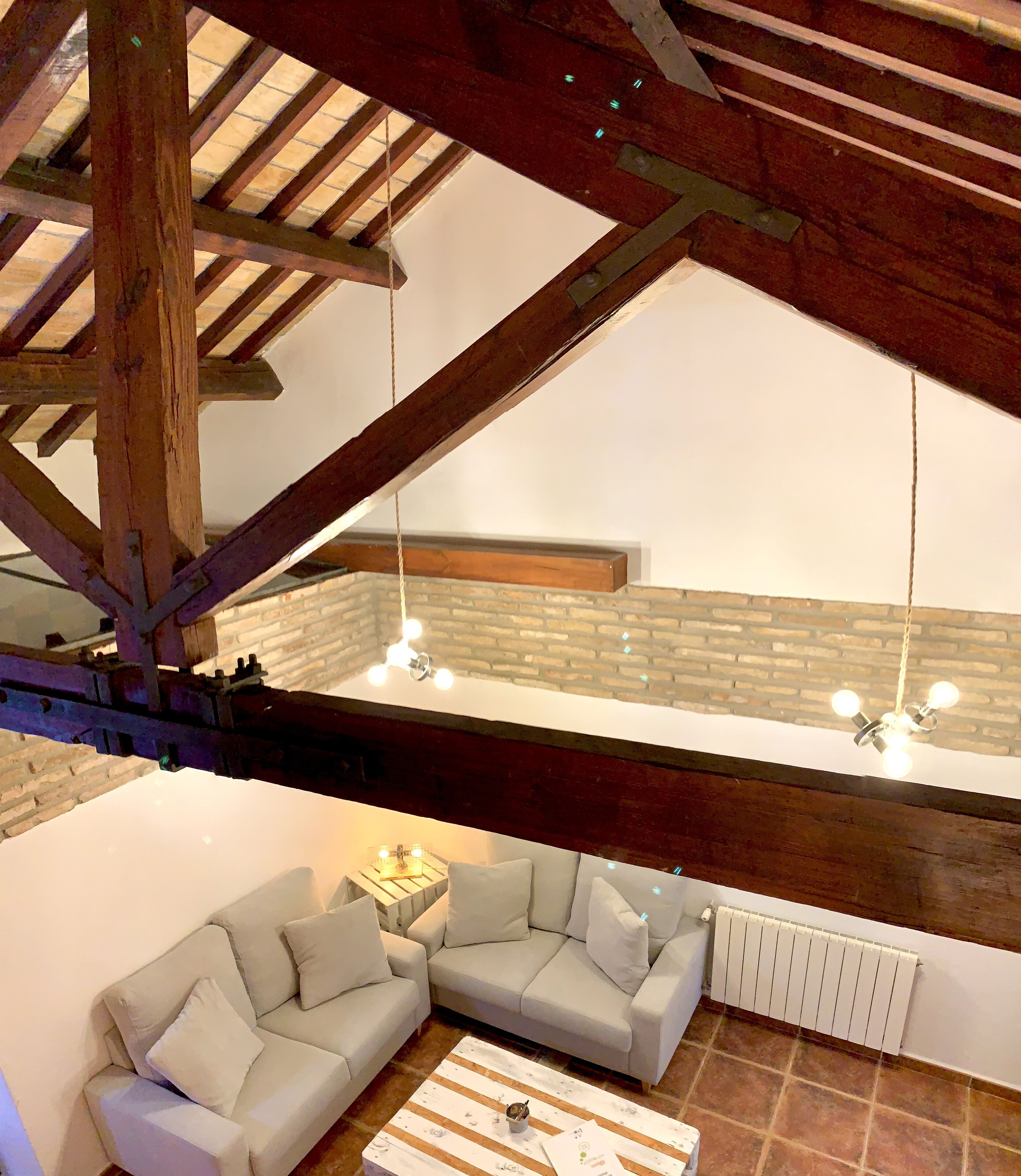 A loft-style room with exposed wooden beams, a white sofa set, a small table with a lamp, and a brick wall.