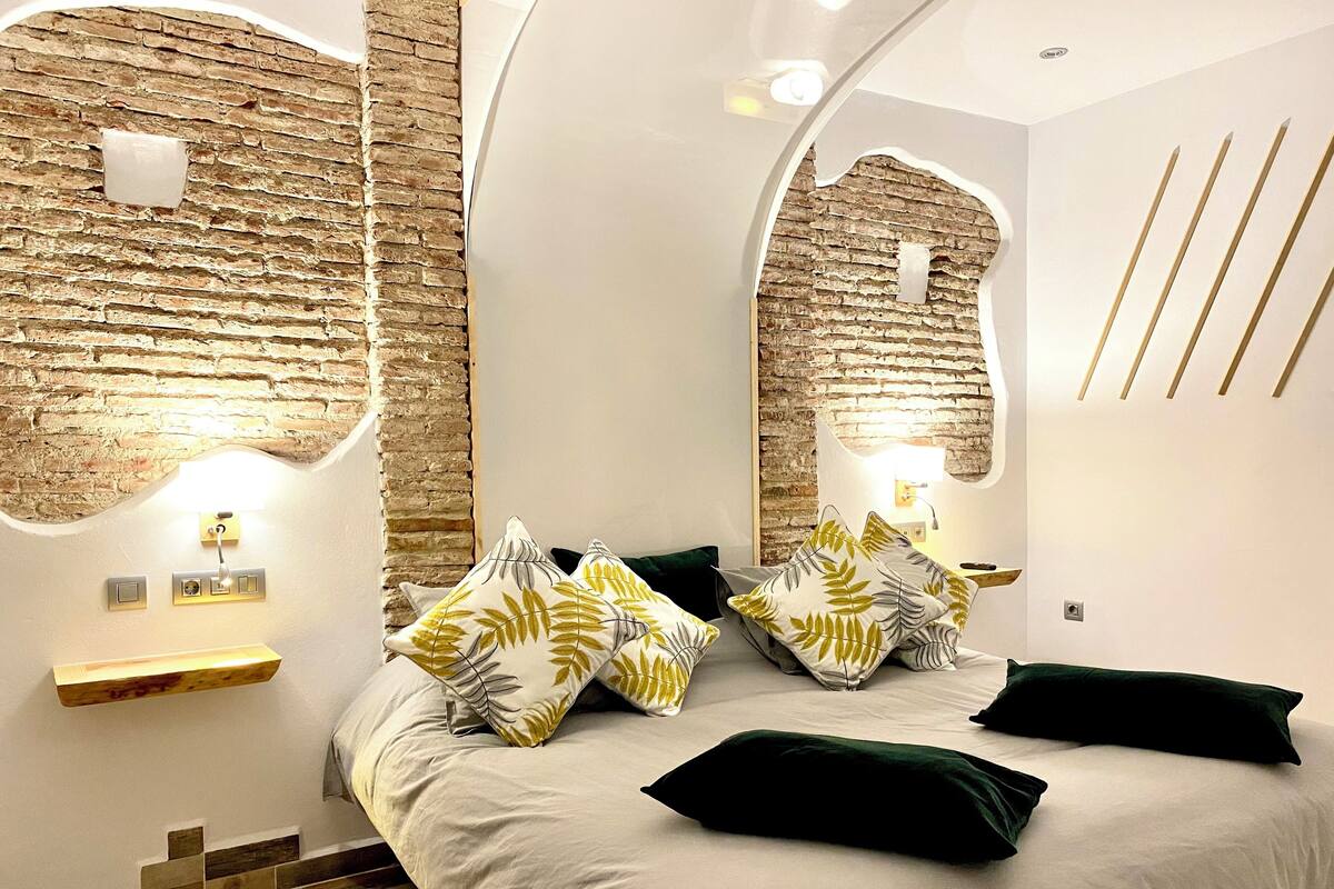 A modern bedroom with a brick wall, a bed with multiple pillows, and a small bedside table.