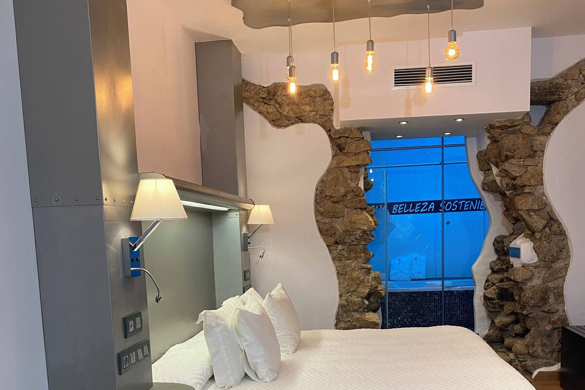 A hotel room with a bed, a sofa, a nightstand, a lamp, a wall-mounted heater, and a view of a pool through a glass wall.