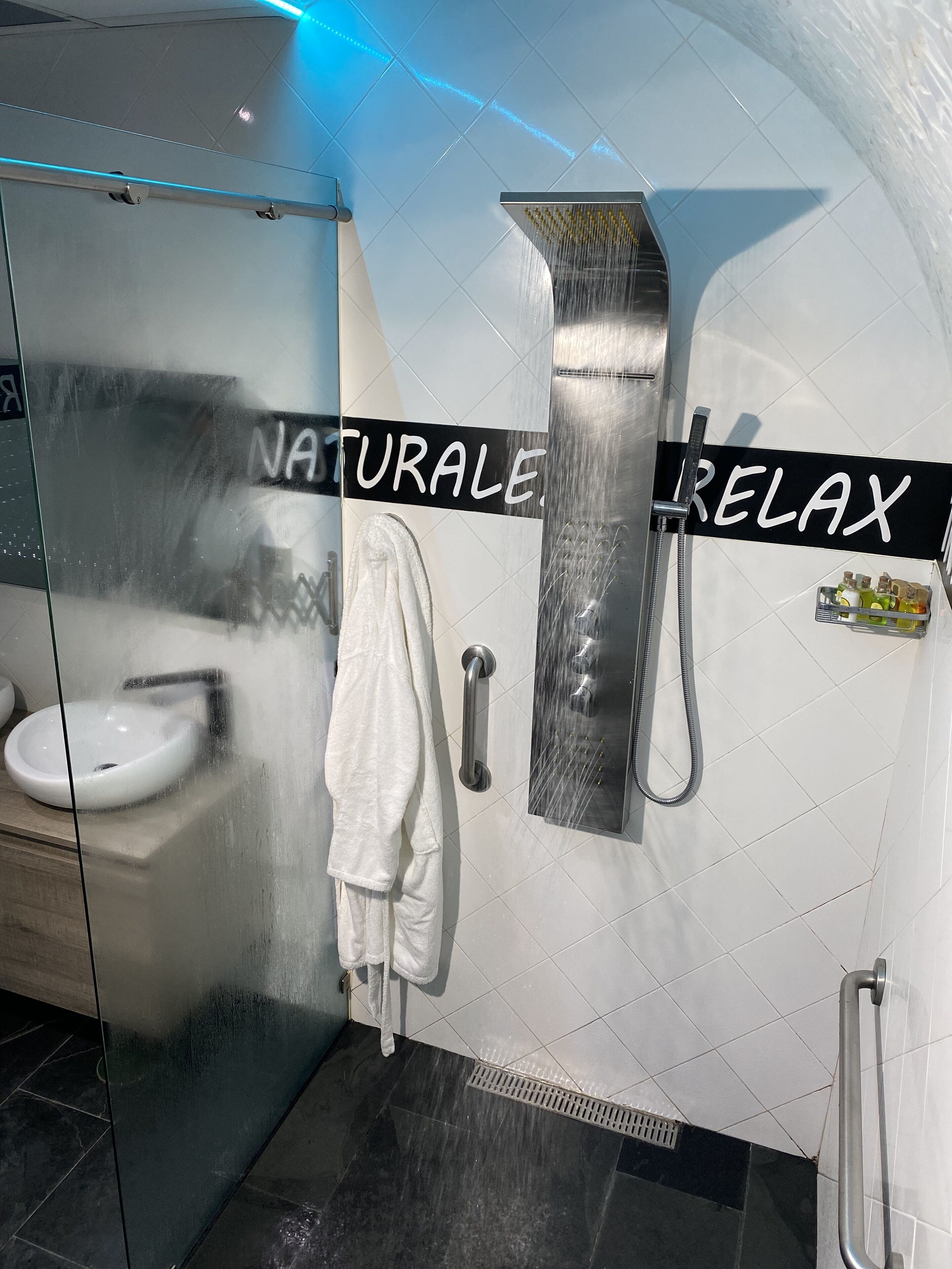 A modern bathroom with a glass shower enclosure, a white sink, a towel rack, and a sign that reads "NATURALE RELAX".